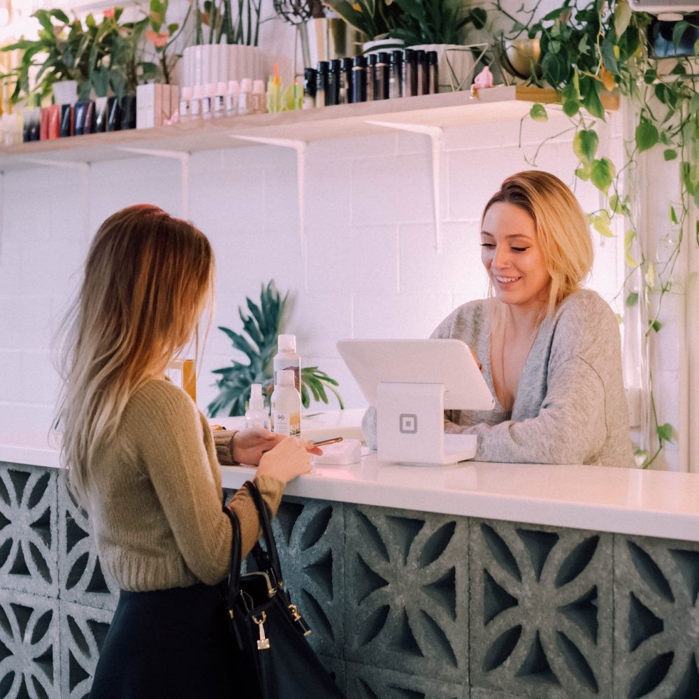 Woman in standing in front of a shelf of beauty products and serving a customer at a till