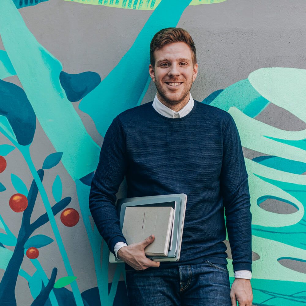 Hombre con una carpeta de notas y una laptop cerrada, apoyado en una pared de colores
